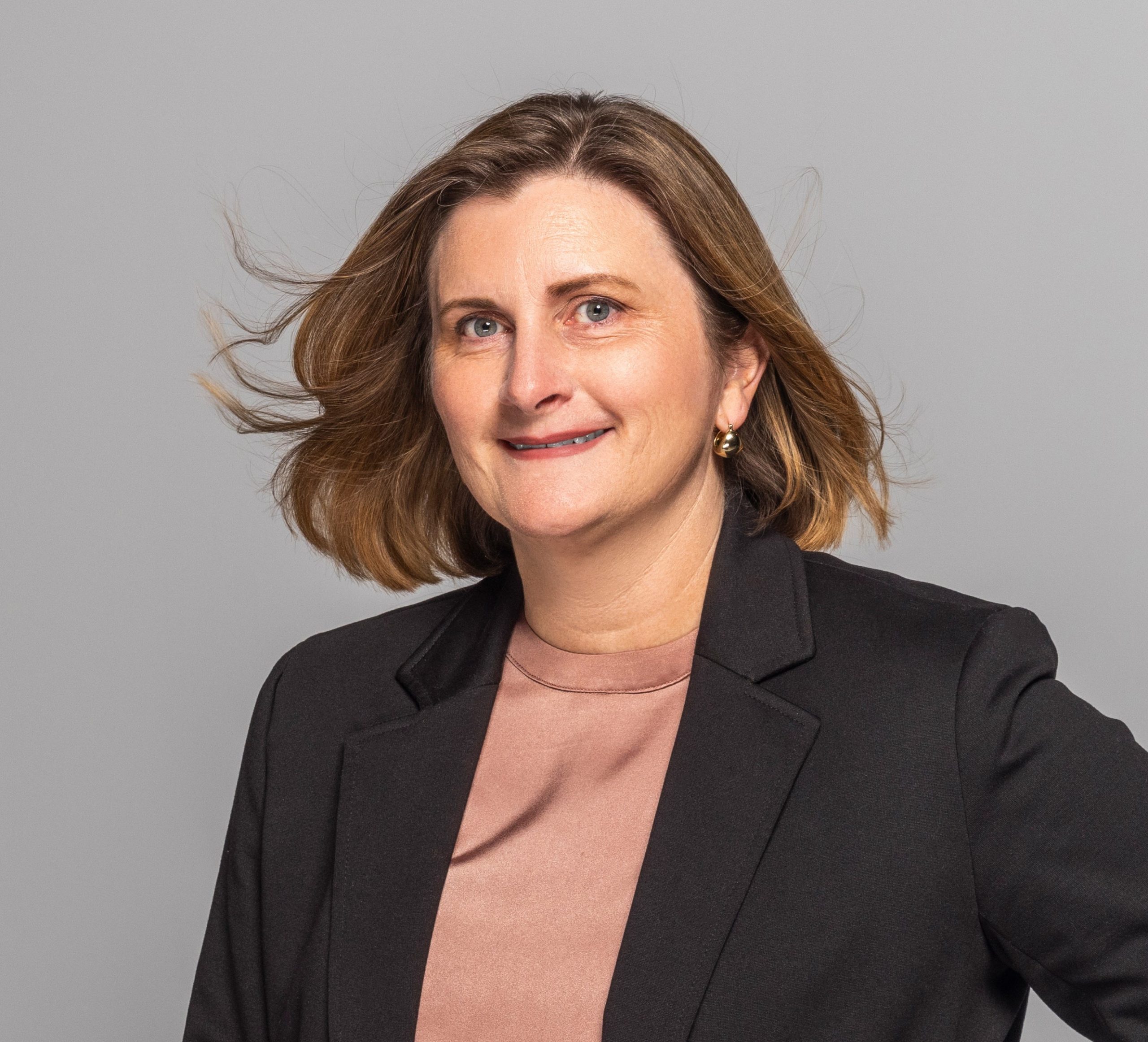 Caucasian woman smiling, wearing coral top and black blazer.